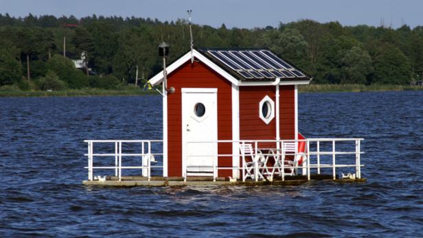 Ein kleines, rotes Haus mit Solaranlage steht auf einer Plattform im Wasser.