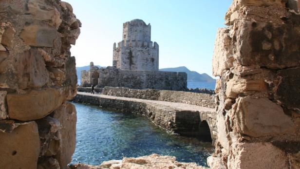 Eine mittelalterliche Burgruine mit einem Turm, verbunden durch einen steinernen Damm mit einem kleinen Durchgang für das Meer.