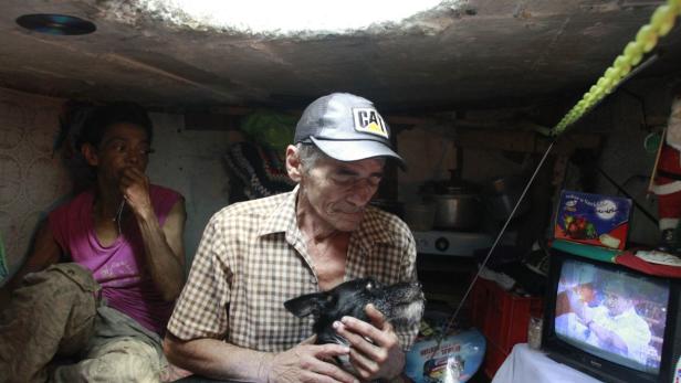 Ein älterer Mann mit Mütze hält einen kleinen schwarzen Hund in einem beengten Raum.