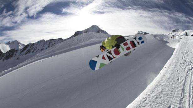 Ein Snowboarder springt mit einem Burton-Board vor einer verschneiten Bergkulisse.
