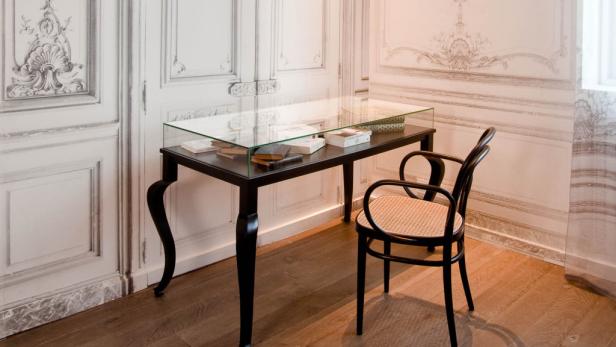 Ein schwarzer Tisch mit Glasplatte und Büchern steht neben einem Stuhl mit Rattansitz.