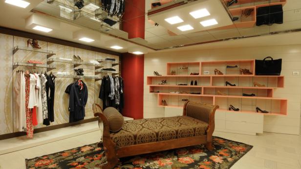 Innenansicht eines Bekleidungsgeschäfts mit Kleidung, Schuhen und einer Ottomane.