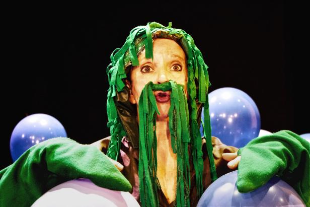 Eine Frau mit grünen Stoffstreifen als Haare und Bart posiert vor blauen Luftballons.