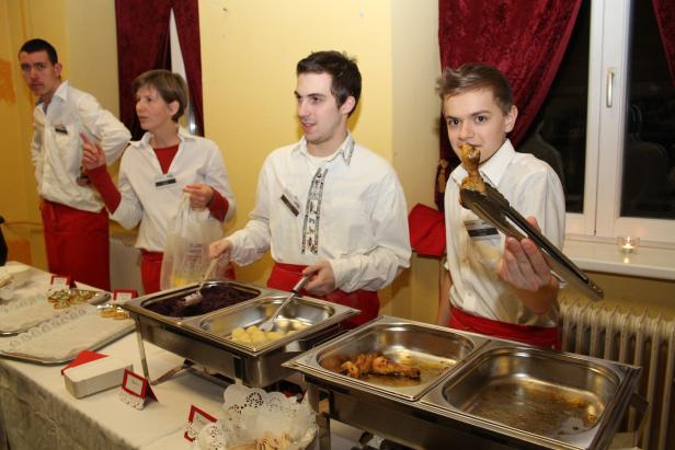 Mehrere Personen stehen an einem Buffet mit Speisen wie Rotkohl, Kartoffeln und Hähnchenkeulen.