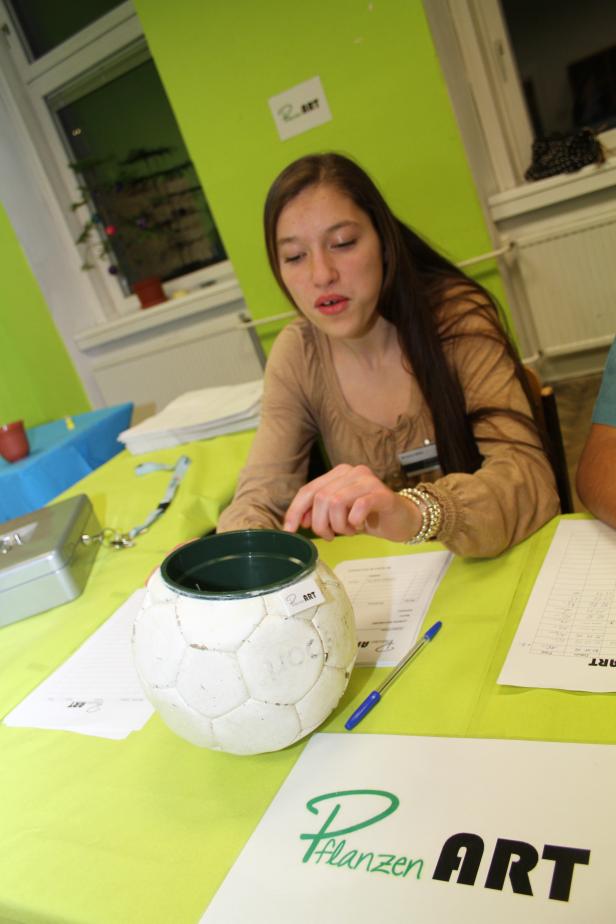 Eine junge Frau sitzt an einem Tisch mit einer Fußball-Spardose und Unterlagen von „Pflanzen ART“.
