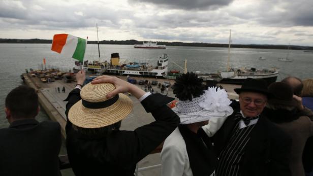 Menschen in historischer Kleidung winken mit einer irischen Flagge einem Schiff zu.