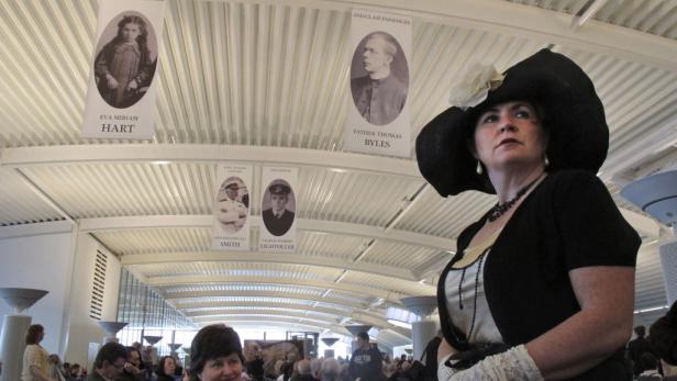 Eine Frau in historischer Kleidung steht unter Bannern mit Porträts von Titanic-Passagieren.
