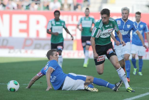 Ein Fußballspieler in Grün versucht, einen am Boden liegenden Spieler in Blau zu überwinden.