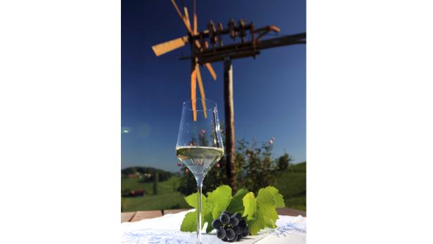 Ein Glas Weißwein und eine Traube vor einer hölzernen Windmühle in einer ländlichen Szene.