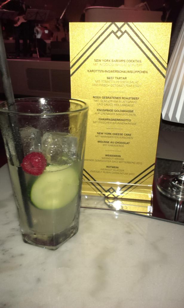 Ein Cocktail mit Eis, Gurke und Himbeere steht vor einer Speisekarte.