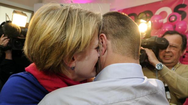 Eine Frau umarmt einen Mann vor einer rosa dekorierten Wand, während sie von Kameraleuten fotografiert werden.