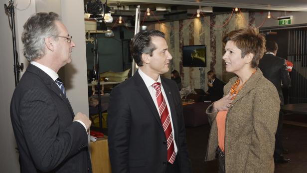 Drei Personen in Anzügen unterhalten sich in einem Raum mit gedämpfter Beleuchtung.