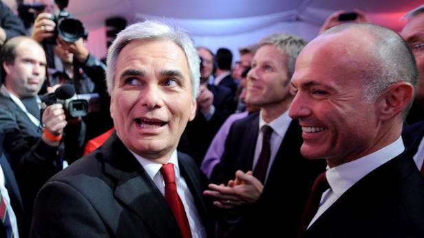 Zwei Männer in Anzügen lächeln, umgeben von Kameras und anderen Personen.