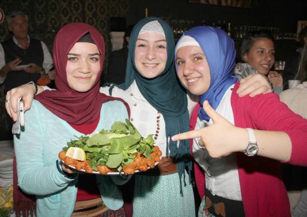 Drei lächelnde junge Frauen mit Kopftüchern präsentieren eine Platte mit türkischen Köfte und Salat.