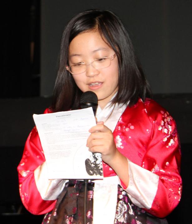 Ein junges Mädchen in traditioneller koreanischer Kleidung liest von einem Blatt Papier in ein Mikrofon.
