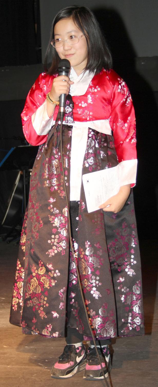 Ein junges Mädchen in traditioneller koreanischer Kleidung hält ein Mikrofon und ein Blatt Papier.