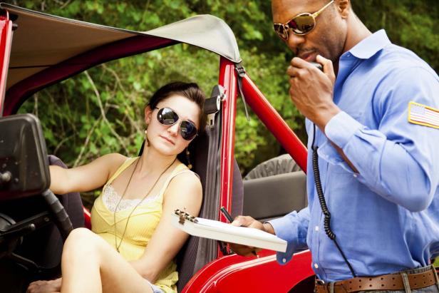 Eine Frau sitzt in einem roten Jeep, während ein Polizist ihr einen Strafzettel ausstellt.