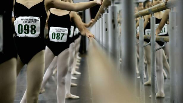 Eine Reihe junger Balletttänzerinnen mit Startnummern an einer Ballettstange.