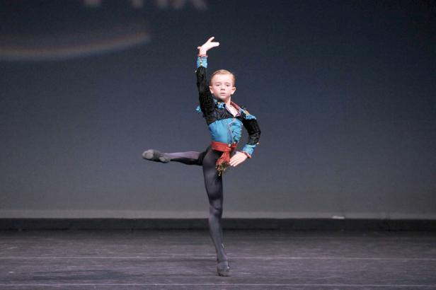 Ein junger Tänzer in einem farbenfrohen Kostüm führt eine Ballettpose auf einer Bühne auf.
