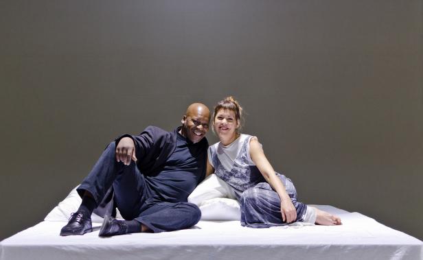 Ein Mann und eine Frau sitzen lächelnd auf einem Bett vor einem grauen Hintergrund.