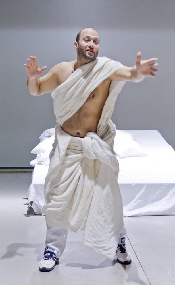 Ein Mann in einer weißen Toga und Turnschuhen steht vor einem weißen Bett.