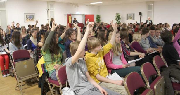 Viele junge Leute sitzen in einem Raum und heben ihre Hände.