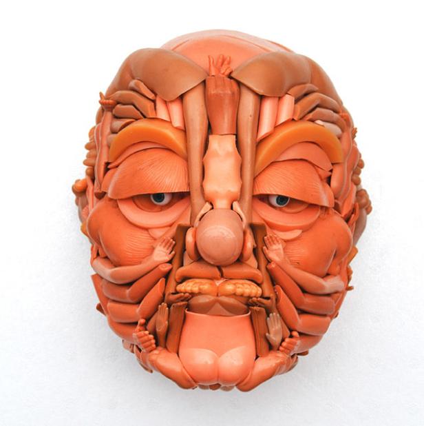 Eine Maske, geformt aus Teilen von Babypuppen, hängt an einer weißen Wand.