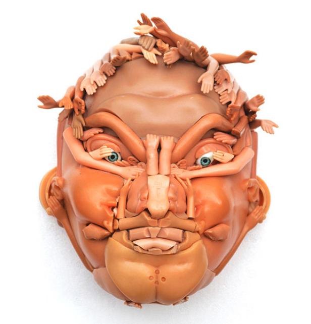 Eine skulpturale Maske, geformt aus Puppenteilen, darunter Hände und Gliedmaßen, auf einem weißen Hintergrund.