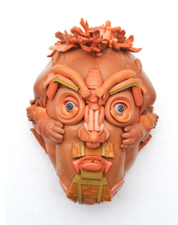 Eine skulpturale Maske aus orangefarbenem Ton mit Augen und Händen verziert.
