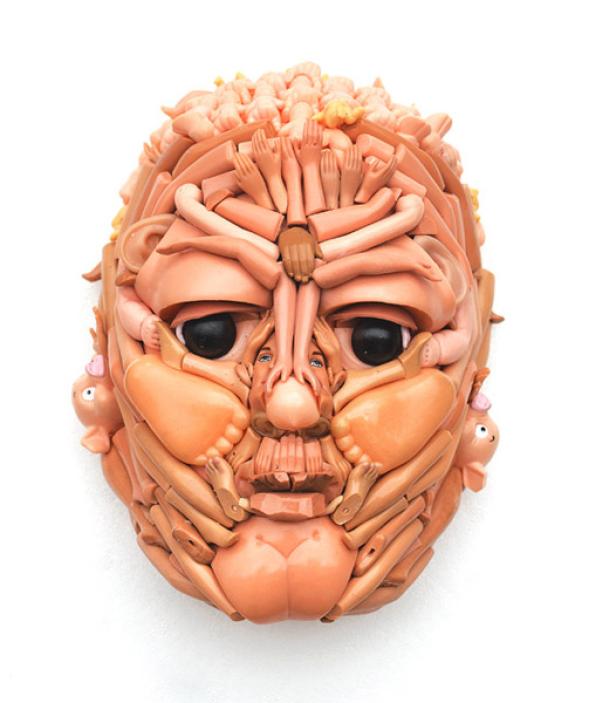 Eine skulpturale Maske, geformt aus Puppenteilen, darunter Arme, Beine und Gesichter.