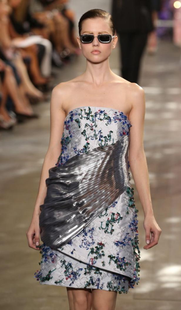 Ein Model mit Sonnenbrille präsentiert ein trägerloses Kleid mit silbernen Akzenten auf dem Laufsteg.