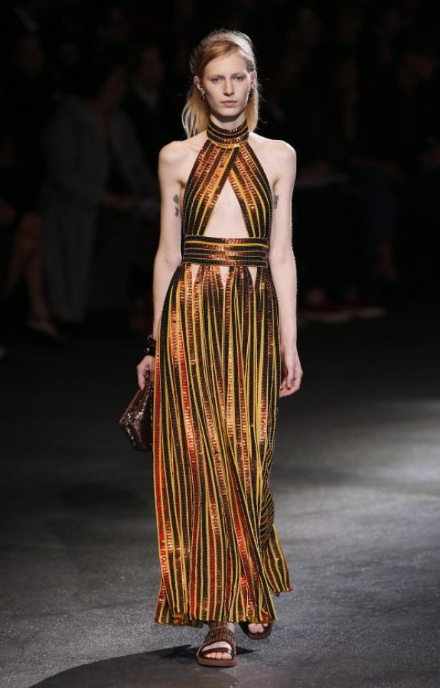 Ein Model präsentiert ein gestreiftes, gelb-schwarz-rotes Kleid mit Cut-Out auf einem Laufsteg.