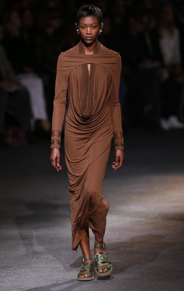 Eine Frau in einem braunen Kleid und grünen Sandalen auf einem Laufsteg.