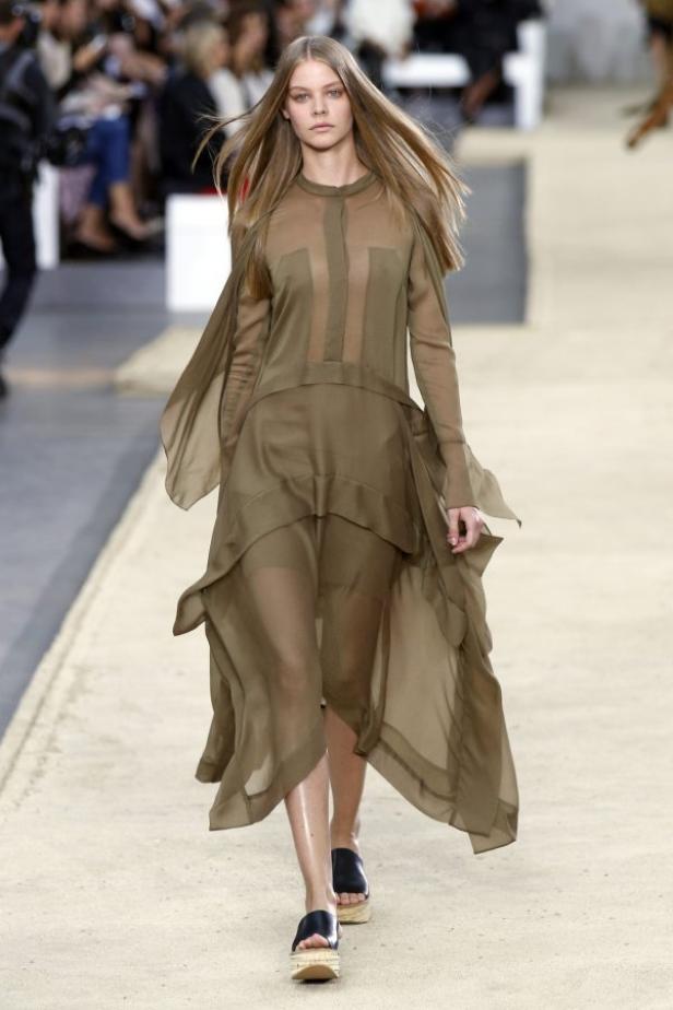 Ein Model präsentiert ein transparentes, khakifarbenes Kleid auf dem Laufsteg.