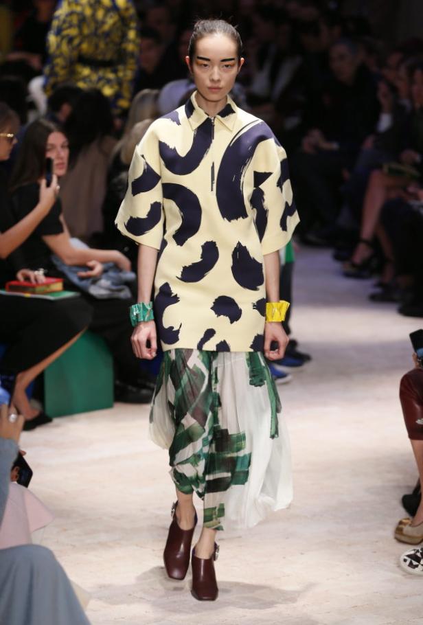 Eine Frau präsentiert ein gelbes Kleid mit dunklen Pinselstrichen und einen grün-weißen Rock auf dem Laufsteg.