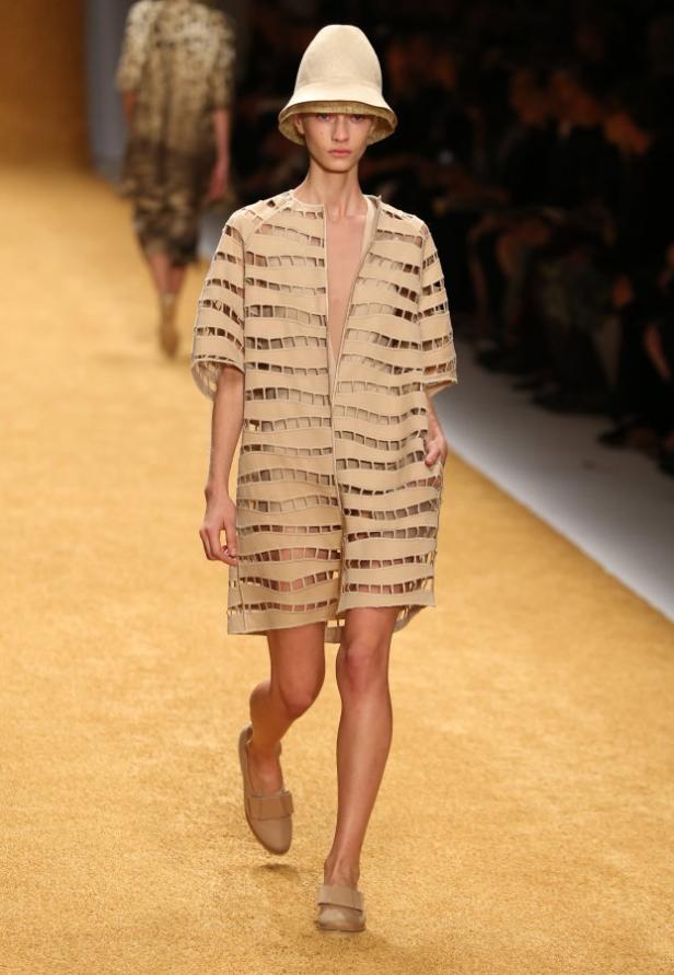 Ein Model auf dem Laufsteg präsentiert ein beiges Kleid mit quadratischen Ausschnitten und einen passenden Hut.