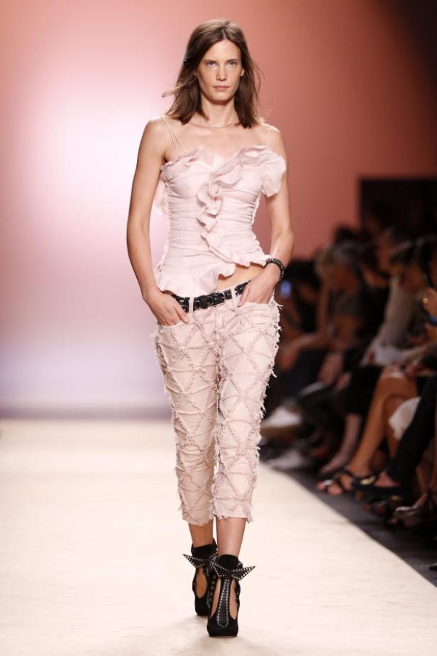 Ein Model in einem rosa Outfit mit Rüschenoberteil und Jeans auf einem Laufsteg.