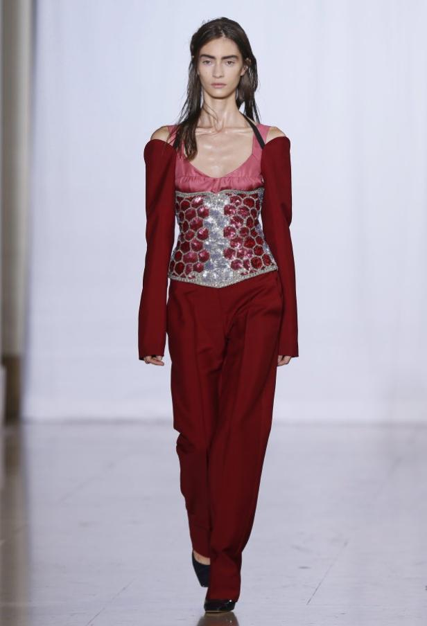 Eine Frau in einem roten Hosenanzug mit einem verzierten Oberteil auf einem Laufsteg.