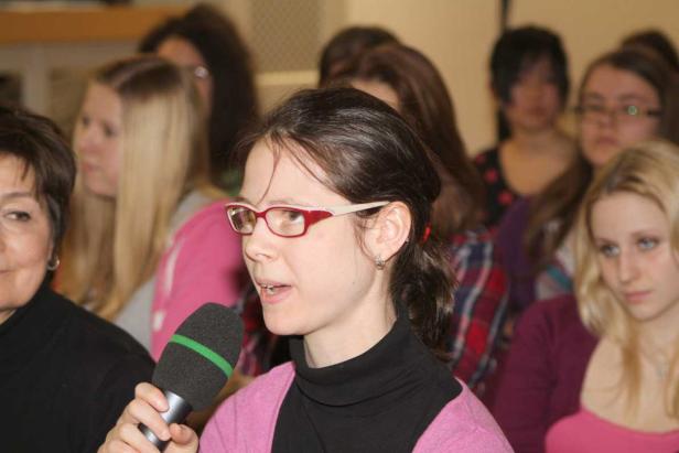 Eine junge Frau spricht in ein Mikrofon vor einer Gruppe von Zuhörern.