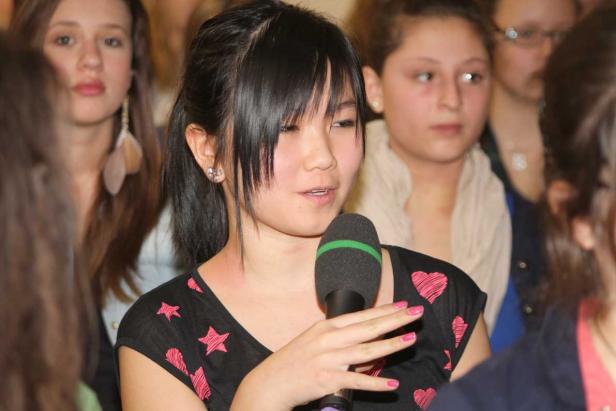 Eine junge Frau spricht in ein Mikrofon vor einer Gruppe von Menschen.