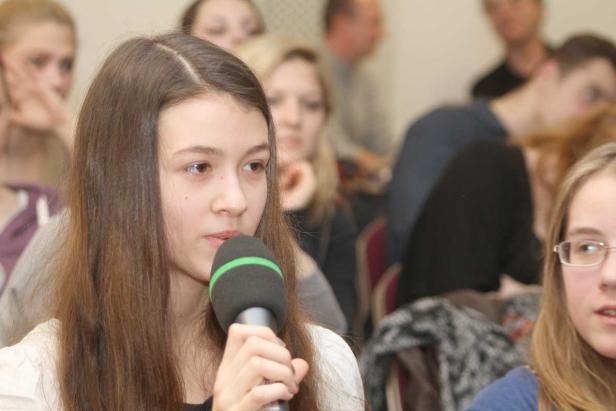 Ein junges Mädchen spricht in ein Mikrofon vor einer Gruppe von Zuhörern.
