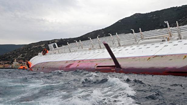 Ein gekentertes Schiff liegt auf der Seite im Wasser nahe einer Küstenstadt.