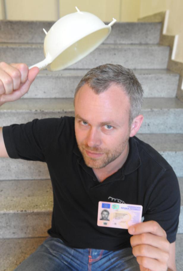 Ein Mann mit einem Nudelsieb auf dem Kopf hält einen Führerschein in der Hand.