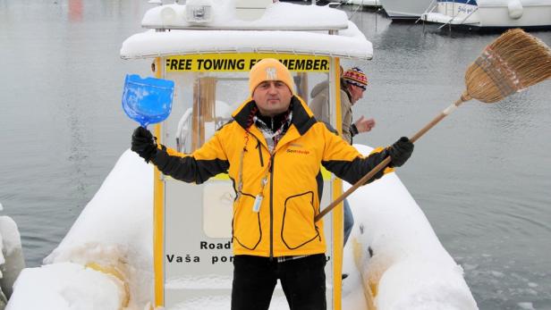 Ein Mann auf einem schneebedeckten Boot hält eine blaue Schaufel und einen Besen.