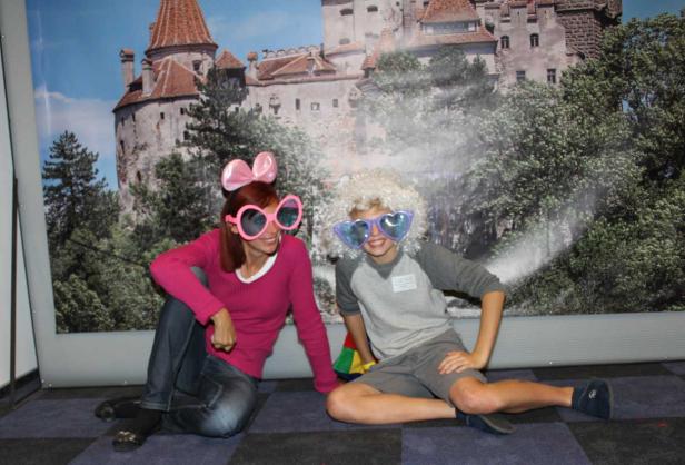 Zwei Personen mit übergroßen Brillen und Perücken posieren vor einem Bild des Schlosses Bran.