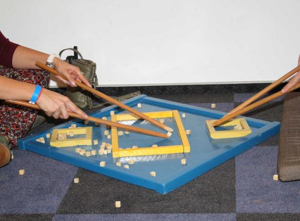 Personen spielen mit Stäben und kleinen Holzklötzen auf einem blauen Brett.