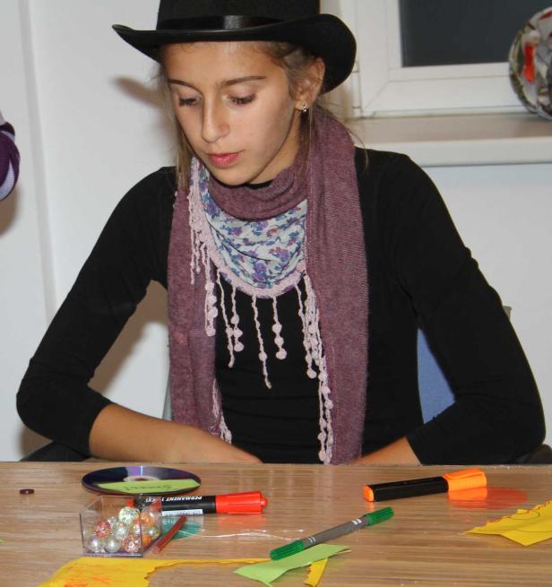 Ein Mädchen mit Hut und Schal sitzt an einem Tisch mit Bastelmaterialien.