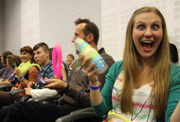 Eine aufgeregte Frau hält eine bunte Flasche in einem Raum voller Menschen.