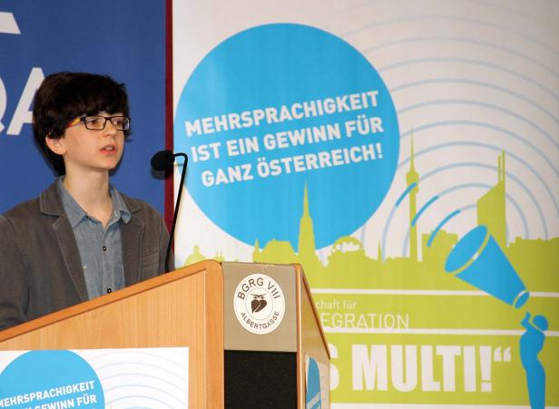 Ein junger Redner steht an einem Pult mit dem Slogan „Mehrsprachigkeit ist ein Gewinn für ganz Österreich!“.