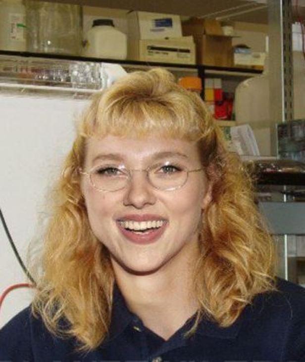 Eine lächelnde Frau mit blonden, gewellten Haaren und Brille steht in einem Labor.